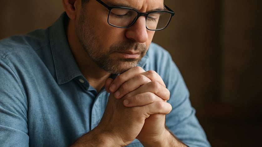 Homem de óculos e camisa azul, de olhos fechados, com as mãos unidas diante de um livro aberto.