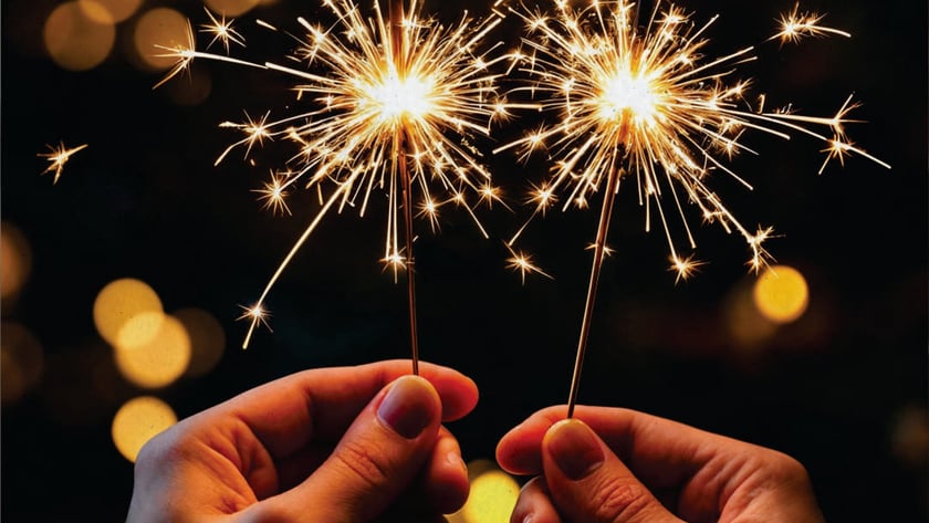 Duas mãos seguram estrelinhas acesas, soltando faíscas douradas à noite.