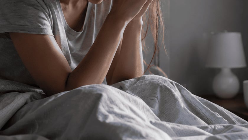 Mulher de cabelos longos sentada na cama, com a cabeça apoiada nas mãos unidas.