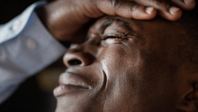 Homem de olhos fechados, com lágrimas no rosto, apoia a mão na testa.