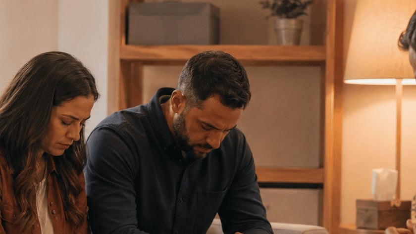 Dois homens sentados, de cabeça baixa e mãos unidas, rezam numa sala com uma Bíblia aberta.