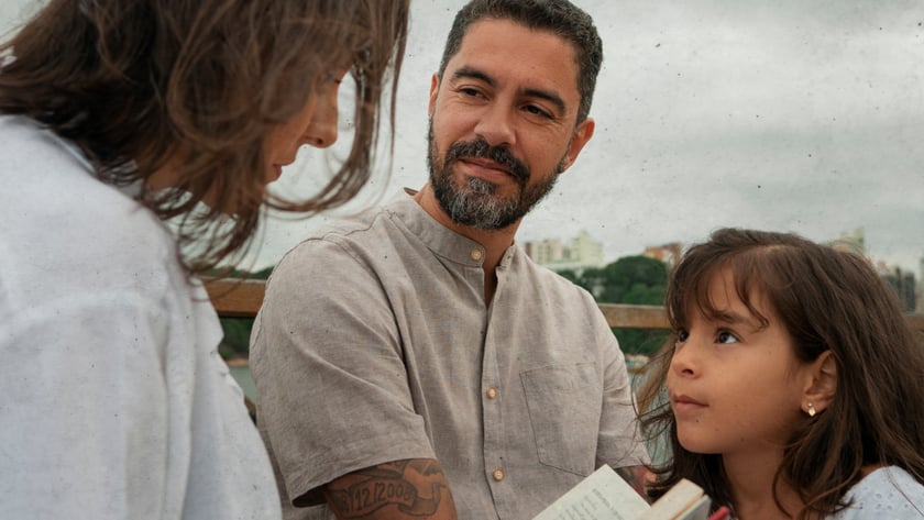 Homem de barba, mulher e menina conversam ao ar livre segurando um livro aberto.