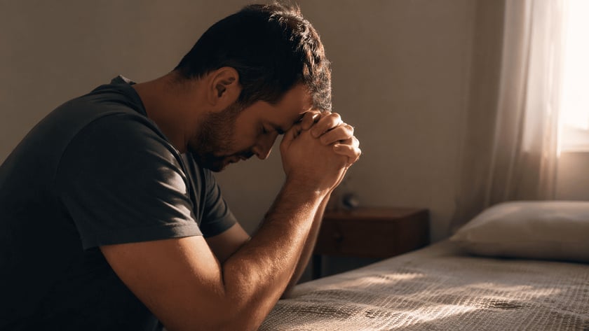 Homem de camiseta cinza, de olhos fechados e mãos postas na testa, ao lado de uma cama.