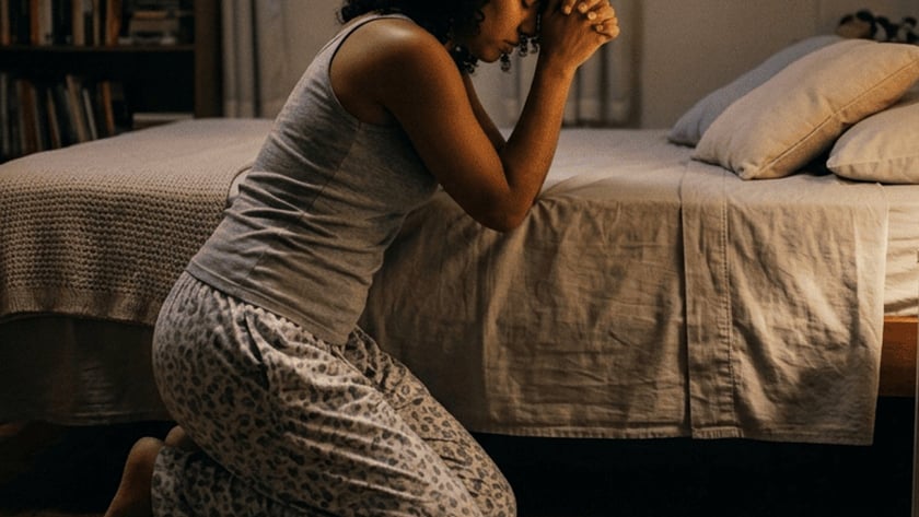 Mulher de pijama ajoelhada ao lado da cama, com as mãos unidas, rezando no quarto.