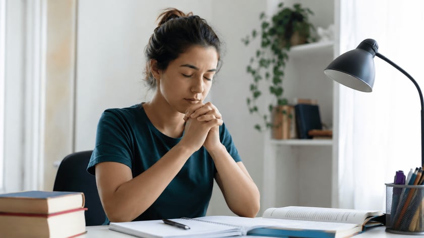 Mulher de olhos fechados e mãos unidas à mesa de estudos, com livros abertos.
