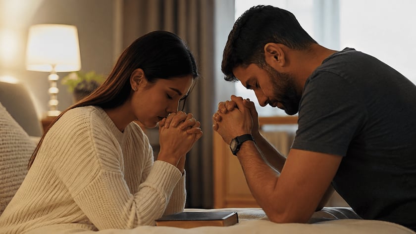 Homem e mulher de mãos unidas e cabeças baixas, rezando ao lado de uma cama com um livro.