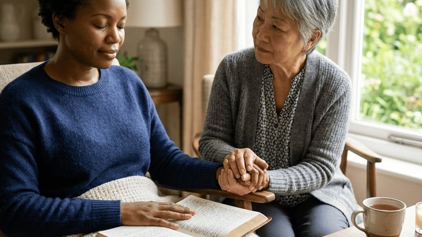 Duas mulheres sentadas de mãos dadas, uma com uma Bíblia aberta no colo, numa sala.
