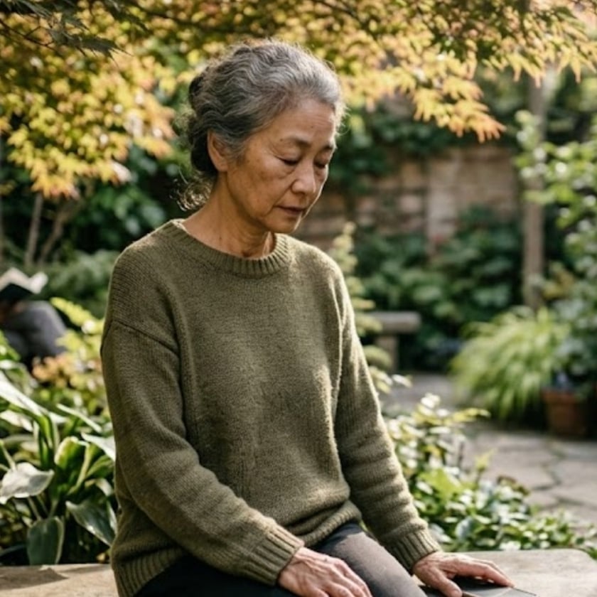 Mulher idosa de cabelo grisalho e suéter verde senta de olhos fechados em um jardim.