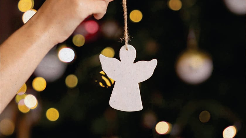 Mão segura enfeite de anjo branco pendurado por barbante diante de árvore de Natal.