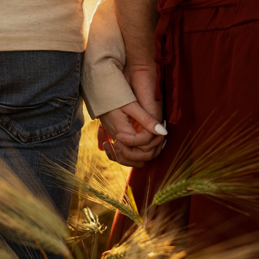 Duas pessoas de mãos dadas em um campo de trigo, uma de jeans e outra de vestido vermelho.