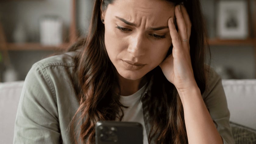 Mulher de cabelo escuro sentada no sofá, olhando o celular com a mão na testa.