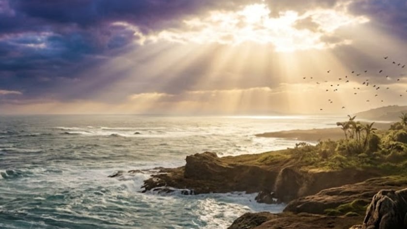 Costa rochosa com vegetação tropical, mar agitado e raios de sol entre nuvens escuras.