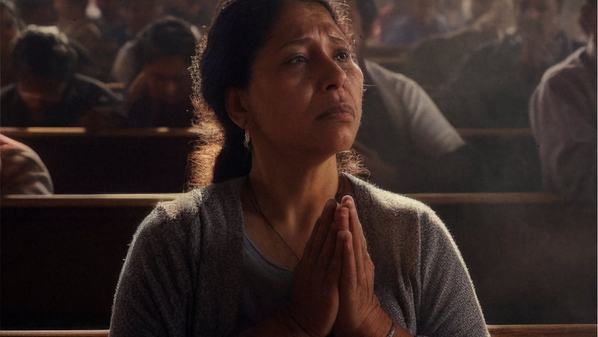 Mulher de cardigã cinza reza de mãos juntas em um banco de igreja lotada.