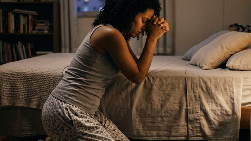 Mulher de pijama ajoelhada ao lado da cama, com as mãos unidas, rezando no quarto.