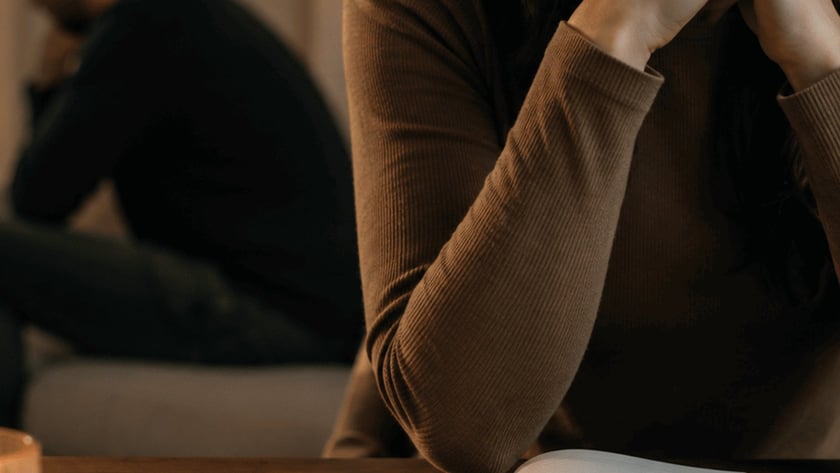 Mulher de mãos unidas diante de um caderno aberto, com homem sentado ao fundo.