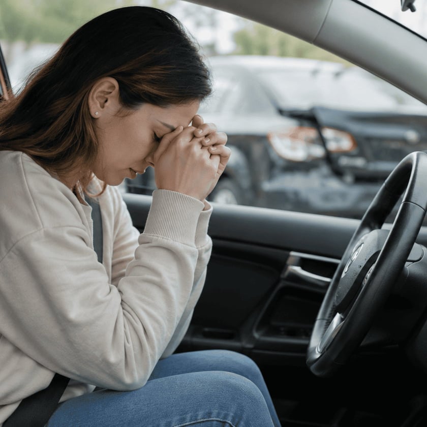 Mulher sentada ao volante de um carro, com o rosto apoiado nas mãos unidas.