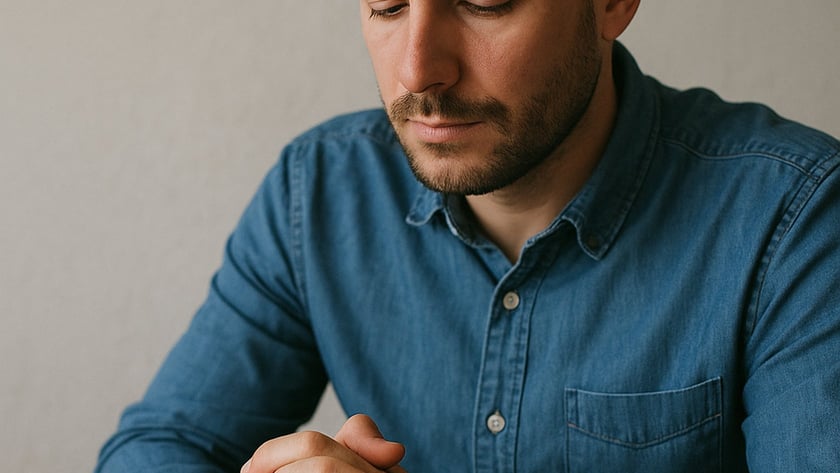 Homem de barba curta e camisa jeans, sentado à mesa, olha para as mãos entrelaçadas.