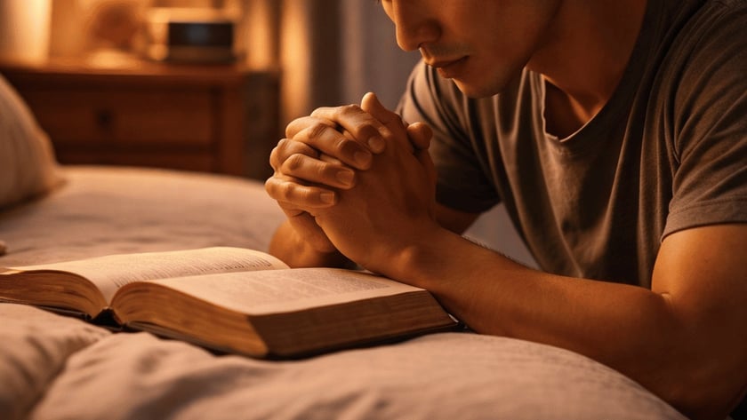 Homem de olhos fechados e mãos juntas ora ao lado de uma Bíblia aberta na cama.