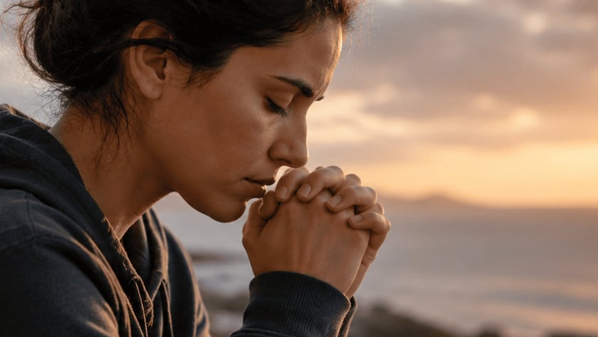 Mulher de olhos fechados e mãos unidas junto ao rosto, à beira-mar, ao entardecer.