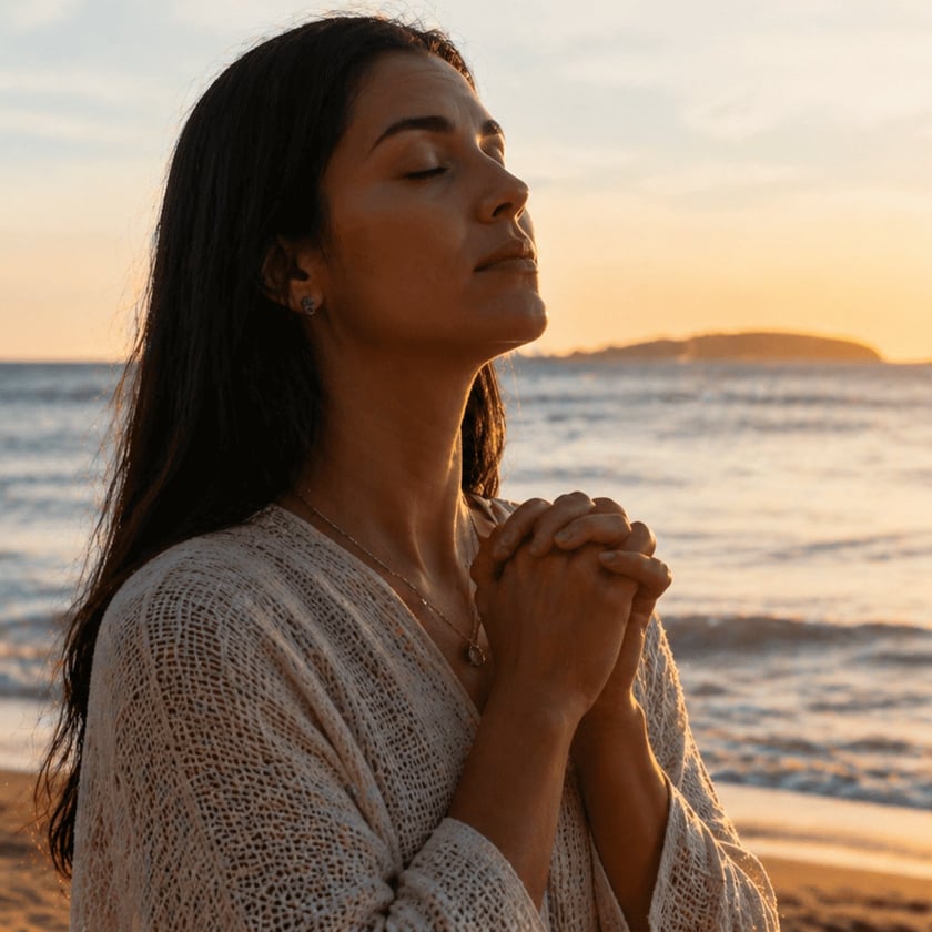 Mulher de cabelos escuros e olhos fechados, com as mãos unidas, na praia.