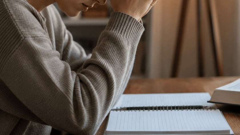 Mulher de cabeça baixa e mãos unidas à testa, à mesa com livro, caderno e caneca.