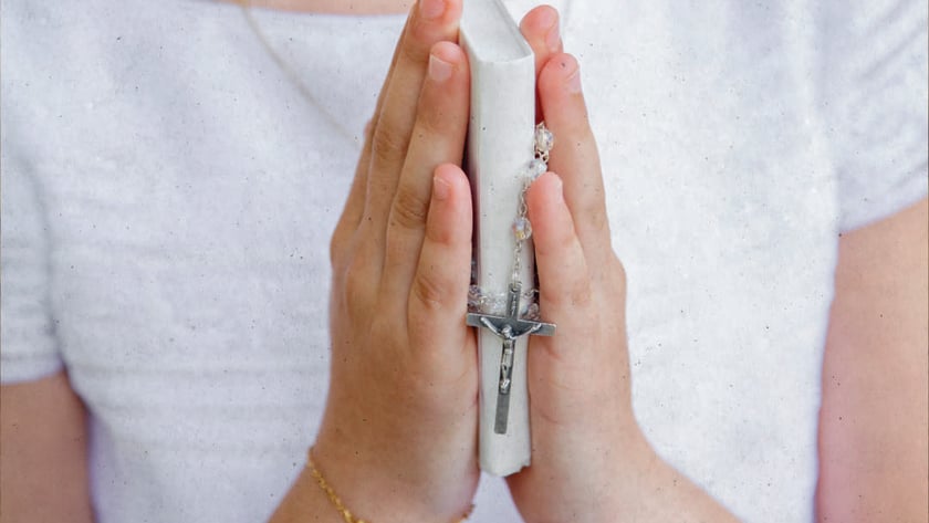Criança de blusa branca segura as mãos postas com um livro branco e um terço.