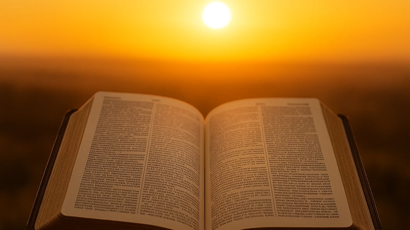 Duas mãos seguram uma Bíblia aberta diante do sol se pondo no horizonte.