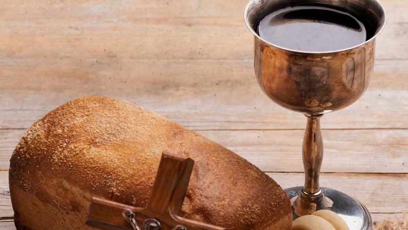 Crucifixo de madeira apoiado em pão de forma, ao lado de cálice com vinho e pãezinhos.