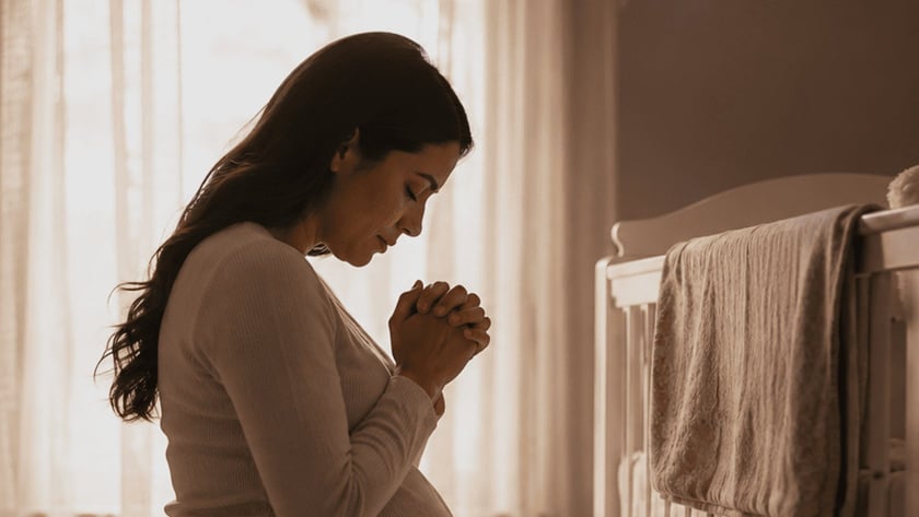 Mulher grávida ajoelhada com as mãos unidas, de olhos fechados, em um quarto de bebê.