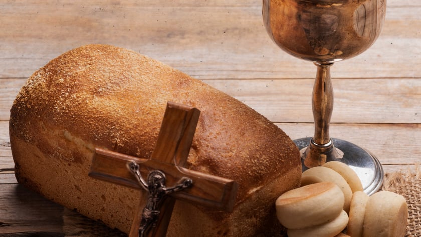 Crucifixo de madeira apoiado em pão de forma, ao lado de cálice com vinho e pãezinhos.