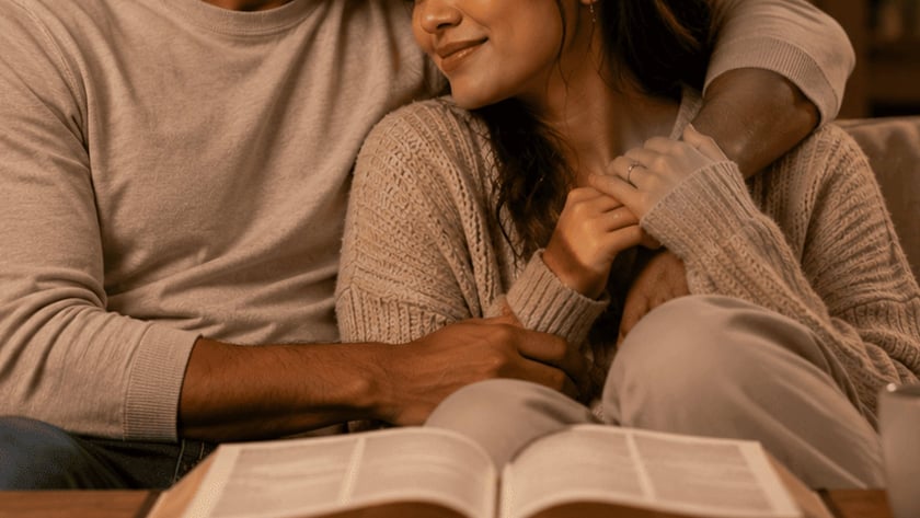 Homem beija a testa de uma mulher abraçada a ele no sofá, com vela e livro.