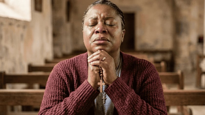 Mulher de cardigã vinho reza com um terço nas mãos em um banco de igreja.