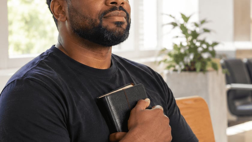 Homem de barba e camiseta preta sentado, segurando uma Bíblia junto ao peito.