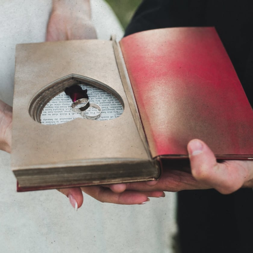 Duas pessoas seguram um livro aberto com recorte em coração contendo duas alianças de prata.