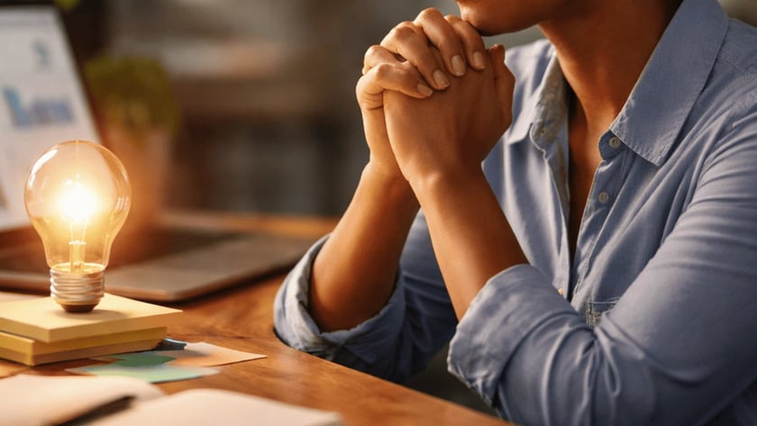 Mulher de olhos fechados e mãos unidas à mesa, com lâmpada acesa e notebook com gráficos.