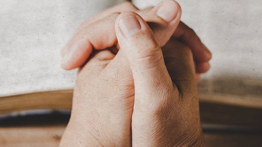 Mãos postas em oração sobre uma Bíblia aberta apoiada em uma mesa de madeira.