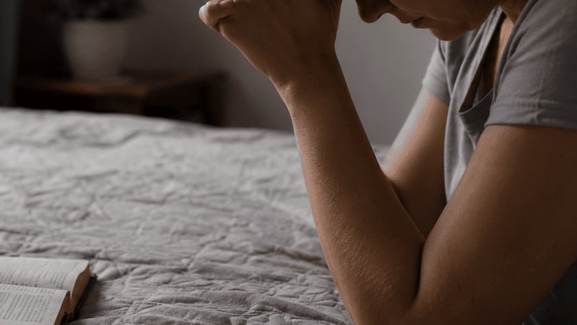 Mulher de cabeça baixa e mãos unidas ao lado de uma Bíblia aberta sobre a cama.