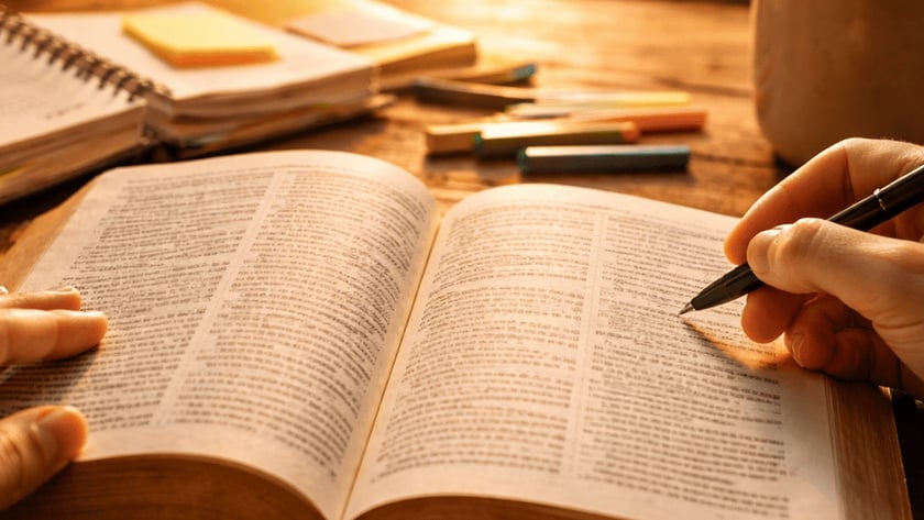 Mãos de uma pessoa sobre um livro aberto, com caneta, cadernos e xícara na mesa.