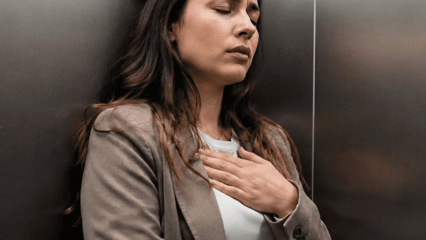 Mulher de blazer marrom encostada na parede de um elevador, com a mão no peito.