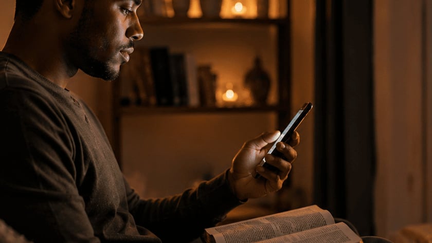 Homem sentado olha o celular, com uma Bíblia aberta no colo, em sala com velas.