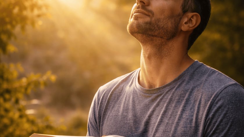 Homem de camiseta cinza, com a cabeça erguida e olhos fechados, segura um livro aberto ao ar livre.