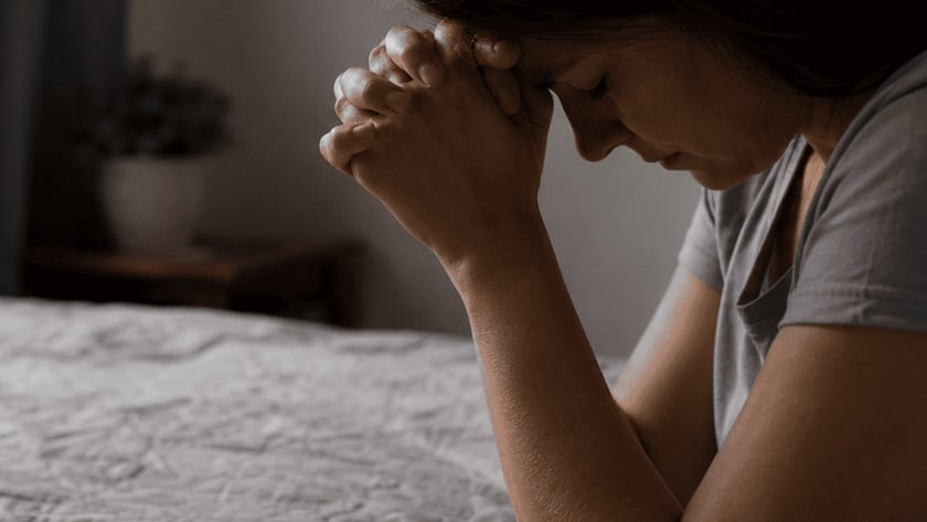 Mulher de cabeça baixa e mãos unidas ao lado de uma Bíblia aberta sobre a cama.