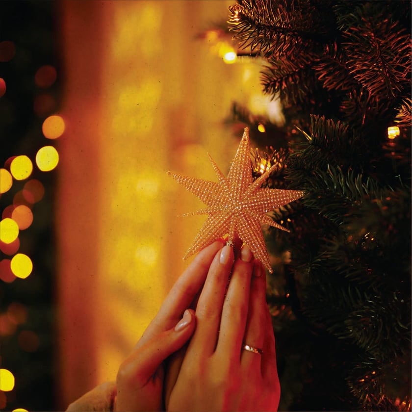 Duas mãos seguram uma estrela dourada de contas junto a uma árvore de Natal.