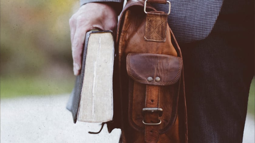 Homem de paletó segura um livro grosso e carrega uma bolsa de couro marrom.