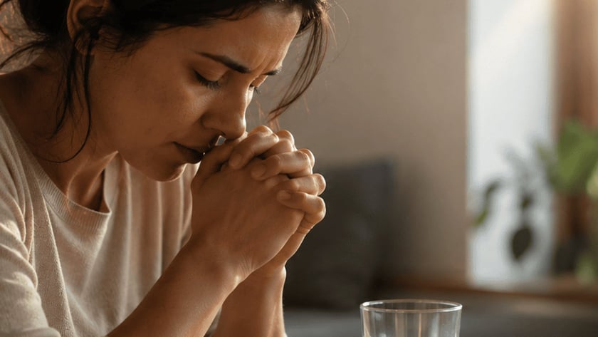 Mulher de olhos fechados e mãos postas, sentada à mesa ao lado de um copo d'água.