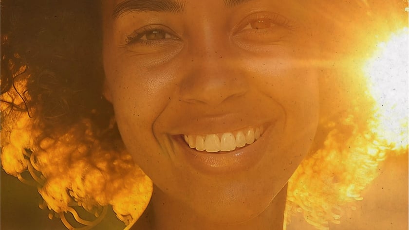 Mulher de cabelo cacheado sorri para a câmera, com o sol atrás dela.