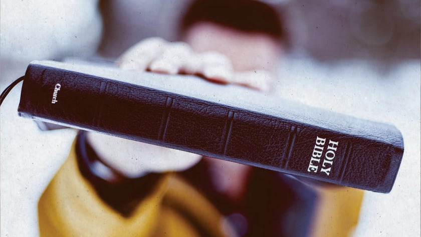 Pessoa de casaco amarelo estende uma Bíblia preta de capa de couro: "HOLY BIBLE".
