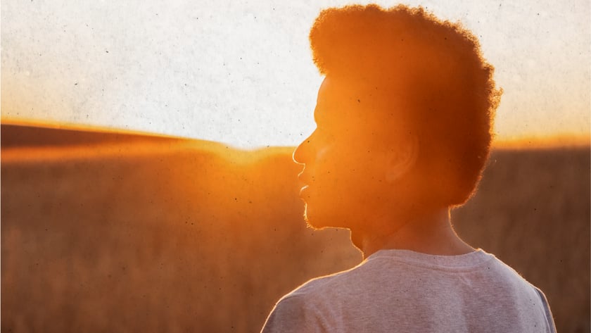 Jovem de camiseta clara e cabelo cacheado olha de perfil para um campo de grama seca.