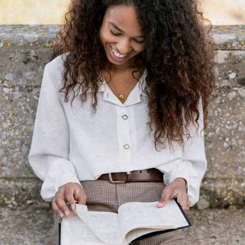 Mulher de cabelo cacheado sorri enquanto lê um livro, sentada junto a um muro de pedra.