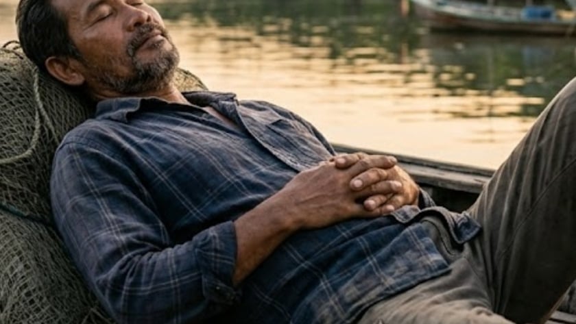 Homem de barba dorme recostado em redes de pesca num barco, perto de um rio.